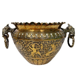 Puede incluir: Jarrón de latón ornamentado con intrincados grabados en relieve. El jarrón presenta un borde festoneado, asas con forma de cabeza de elefante y escenas detalladas de figuras y animales. El color es principalmente dorado con algunos acentos más oscuros.
