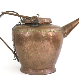 May include: A vintage copper teapot with a hammered finish and a hinged lid. The teapot has a long, curved spout and a handle with a chain attached.