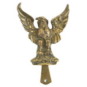 Könnte beinhalten: Ein Türklopfer aus Messing in Form eines Adlers mit ausgebreiteten Flügeln. Der Adler steht auf einem Sockel mit einem dekorativen Design. Der Türklopfer hat einen langen, dünnen Griff.