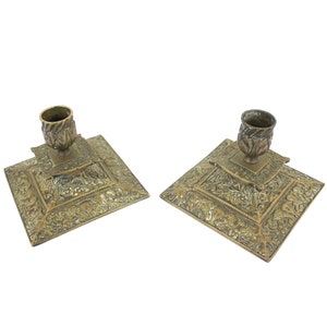Peut inclure: Deux bougeoirs anciens en laiton. Chacun a une base carrée avec des détails floraux ornés et une coupelle centrale pour une bougie. Les bougeoirs sont de couleur laiton vieilli et chaleureux.