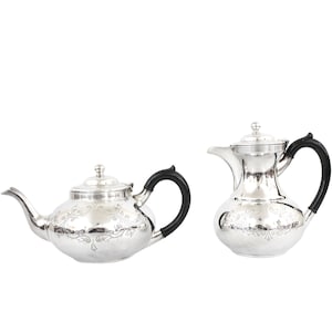Op de afbeelding: Een zilveren theepot en roomkan set met zwarte handvatten en een gegraveerd bloemenmotief.