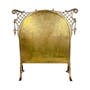 May include: A brass fireplace screen featuring a large, solid, arched central panel with a weathered, golden-toned finish. The frame is adorned with intricate, openwork lattice detailing at the top corners and stands on two curved metal feet.