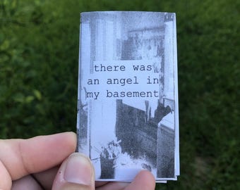 Può includere: Una piccola fanzine rettangolare con una fotografia sgranata in bianco e nero di uno spazio interno. Il testo "there was an angel in my basement" è stampato con un carattere in stile macchina da scrivere al centro della copertina.