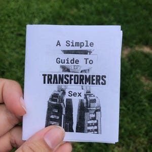 May include: A small white booklet with a black and white printed cover featuring a mechanical robot illustration and the text A Simple Guide To TRANSFORMERS Sex. The booklet is constructed from folded paper with a clean, minimalist aesthetic.