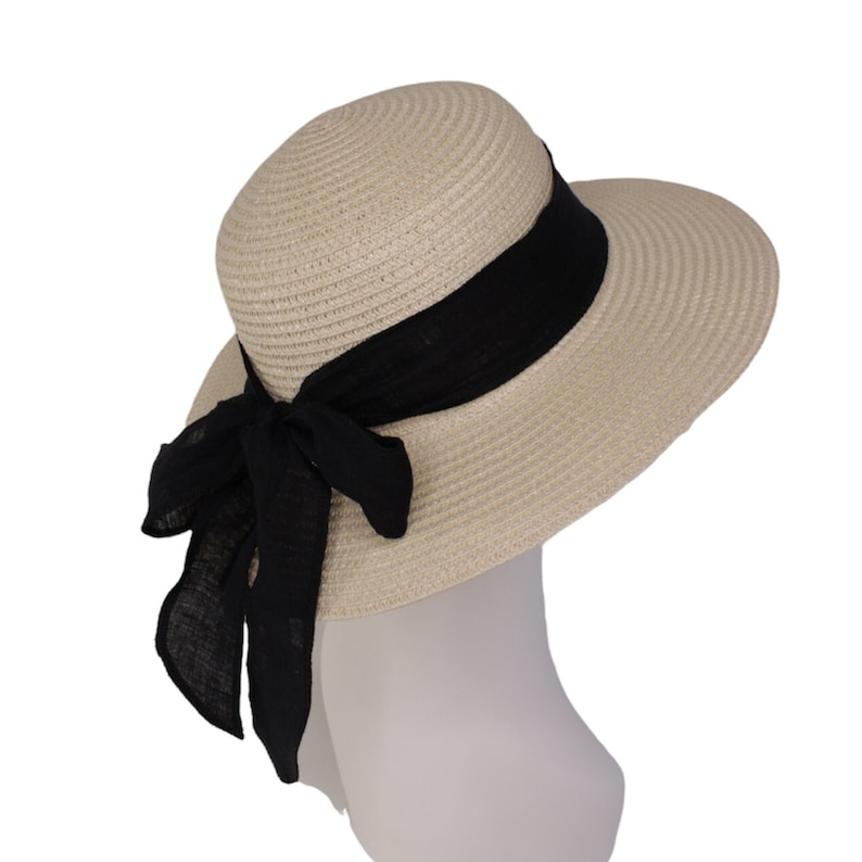 Pu&ograve; includere: Un cappello di paglia beige a tesa larga con un nastro nero legato in un fiocco sul retro.