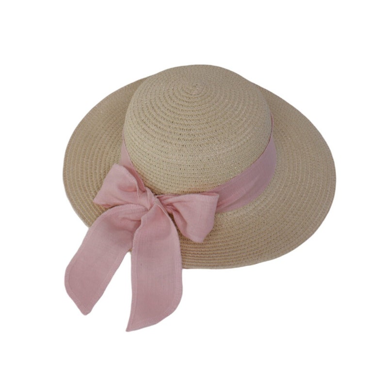 Pu&ograve; includere: Un cappello di paglia a tesa larga con un nastro rosa legato in un fiocco. Il cappello &egrave; beige e ha una trama intrecciata.