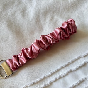 May include: A scrunchie-style wristlet in a dusty rose color. The fabric is gathered and ruched, attached to a gold-toned metal clasp with a key ring. The wristlet is lying on a white, textured surface.