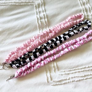 May include: Three fabric lanyards with a gathered, scrunchie-style texture. One lanyard is solid light pink, one features a black and white checkered pattern, and the third is a light pink and white tie-dye pattern. Each includes a silver metal ring.