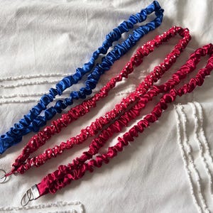 May include: Three satin lanyards in blue, red with white stars, and solid red. Each lanyard has a metal clasp and is laying on a white textured surface. The lanyards are made of a gathered, ruched fabric.