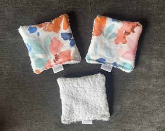 May include: Three white cotton washcloths with a floral pattern and a white terrycloth washcloth. The washcloths are all square and have a frayed edge. The floral washcloths have a blue, pink, and orange watercolor design. The washcloths are all labeled "Cozy Shop".