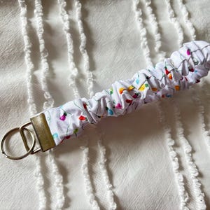May include: A white fabric key fob with a pattern of colorful Christmas lights. The key fob has a metal clasp and two key rings. The fabric is gathered and sewn around a sturdy core.