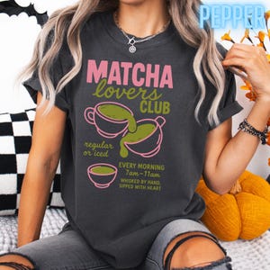 May include: Dark gray t-shirt with the text "MATCHA lovers CLUB" in pink and green. The shirt features a graphic of matcha being poured into a cup, with the words "regular or iced" and "every morning 7am-11am".