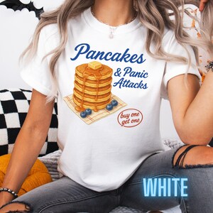 May include: White t-shirt with the text "Pancakes & Panic Attacks" in blue script. The shirt features a graphic of a stack of pancakes with butter and syrup, blueberries, and a "buy one get one" circle.