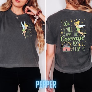 May include: Dark grey t-shirt featuring a cartoon Tinkerbell design on the front and back. The front has Tinkerbell with text, and the back says "We All Have The Courage To Fly" with a castle illustration. The shirt is a comfortable, casual style.