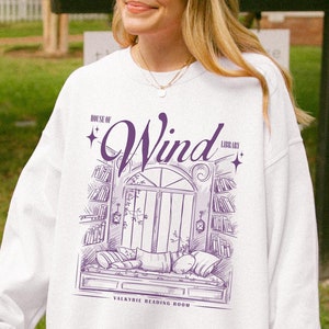 May include: White sweatshirt with a purple graphic of a window with the word "Wind" above it. The graphic also includes the text "Village Reading Room".