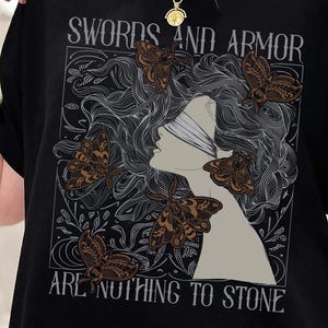 May include: A black t-shirt featuring a graphic print of a silhouette profile surrounded by detailed line-art moths. The text "SWORDS AND ARMOR ARE NOTHING TO STONE" is printed in a serif font above and below the central illustration.