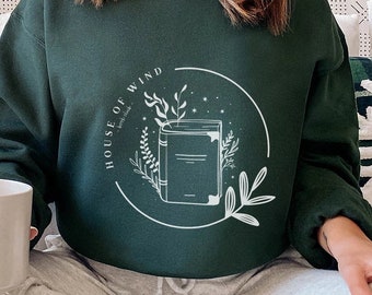 May include: A woman wearing a dark green sweatshirt with the text "HOUSE OF WIND" and a graphic of a book with a floral design.