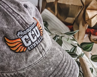 May include: A grey, distressed baseball cap with an orange and yellow embroidered logo that says "Champions CCU Sunball".