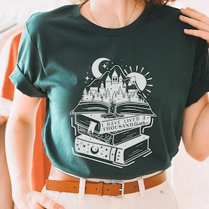 May include: A dark green t-shirt with a white graphic of a castle on a stack of books. The graphic includes a crescent moon, a sun, and the text "I have lived a thousand lives."