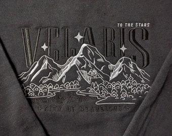May include: Black sweatshirt with white embroidered text that reads "Velaris, City of Starlight" and "To the Stars". The design features a mountain range with a small town at the base.