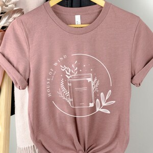 May include: A mauve t-shirt with a white graphic of a book with the text "House of Wind" in a circle.