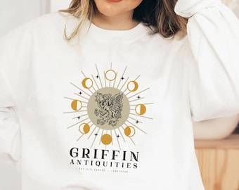 Puede incluir: Sudadera blanca con un gráfico dorado y negro de un grifo con un diseño de luna y estrellas. El texto "Griffin Antiquities" está impreso debajo del gráfico.