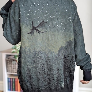 May include: A dark green cardigan with a black silhouette of a dragon flying against a starry night sky. The cardigan has a black knit collar and cuffs.