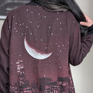 May include: A maroon cardigan with a white crescent moon and stars design. The cardigan features a cityscape silhouette at the bottom.