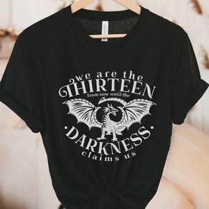 May include: Black t-shirt with a white graphic design featuring a dragon and the text "we are the THIRTEEN from now until the DARKNESS claims us." The shirt is hanging on a wooden hanger.