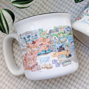 May include: White ceramic mug with a black rim, featuring a colorful map design with text including "Frozen Wastes" and "Gulf of Goro." The mug has a curved handle and is set against a neutral background.