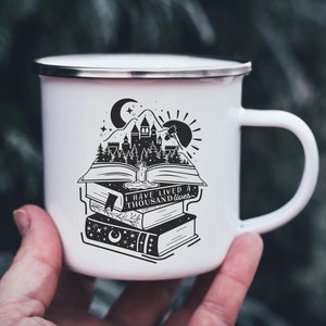 Puede incluir: Taza de esmalte blanco con una ilustración en blanco y negro de un castillo, árboles y un libro con el texto "He vivido mil vidas".