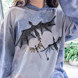 K&ouml;nnte beinhalten: Ein blauer Strick-Kapuzenpullover mit einer Grafik von drei schwarzen Drachen, die vor einem gro&szlig;en, blassen, kreisf&ouml;rmigen Mond fliegen. Der Pullover hat eine texturierte, pixelige Optik mit Farbverl&auml;ufen in Blau, langen &Auml;rmeln und einem gerippten Saum.