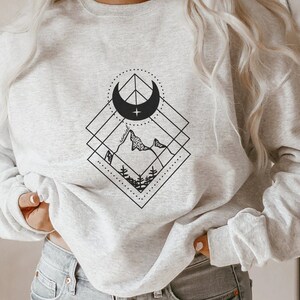 May include: A gray sweatshirt with a black and white graphic design of a crescent moon, mountains, and trees inside a geometric shape.