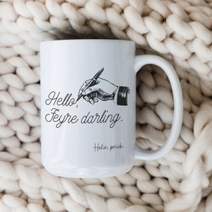 May include: White ceramic mug with a black illustration of a hand holding a pen, with the text "Hello, Feyre darling." and "Hello, prick." in black script. The mug has a large handle.