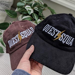 May include: Two corduroy baseball caps with the text "QUEST SQUAD" embroidered in white with a gold lightning bolt. One cap is brown and the other is black.