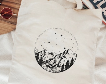 May include: A white tote bag with a black and white illustration of mountains and stars. The text "To the stars who listen and the dreams that are answered" is printed on the bag.
