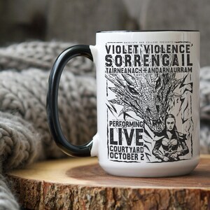 May include: White ceramic mug with a black handle. The mug features a black and white illustration of a dragon and a woman holding a sword. The text on the mug reads "VIOLET VIOLENCE SORRENGAIL TAIRNEANACH+ANDARNAURRAM PERFORMING LIVE COURTYARD OCTOBER 2".