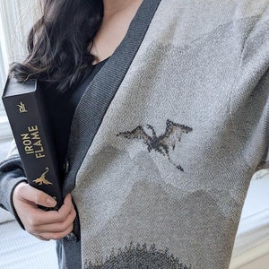 May include: A gray cardigan with a dragon design on the front. The cardigan has a dark gray trim and a button closure. The person is holding a book titled "Iron Flame".