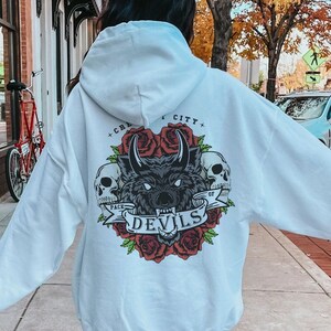 May include: White hooded sweatshirt with a graphic print of a wolf's head with roses and skulls. The text "City of Devils" is printed on the back of the sweatshirt.