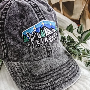 May include: A distressed black baseball cap with a colorful embroidered patch. The patch features a mountain scene with the text "VELARIS CITY OF STARLIGHT". The cap has a curved brim and a worn appearance.