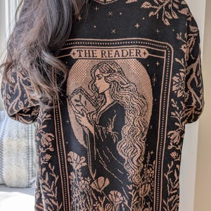 May include: Black sweater featuring a peach-coloured illustration of a woman reading, with floral and star motifs. The text "THE READER" is above the image. The sweater has a crew neck and long sleeves, with a detailed pattern.