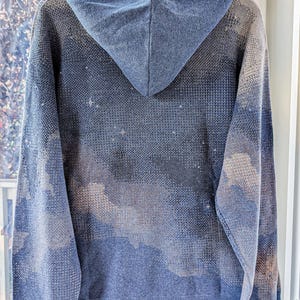K&ouml;nnte beinhalten: Ein blauer Strick-Kapuzenpullover mit einem dezenten, gesprenkelten Muster aus hellen und dunklen T&ouml;nen, das an einen sternenklaren Nachthimmel oder Wolken erinnert. Das Kleidungsst&uuml;ck hat eine Kapuze, lange &Auml;rmel und einen gerippten Saum.