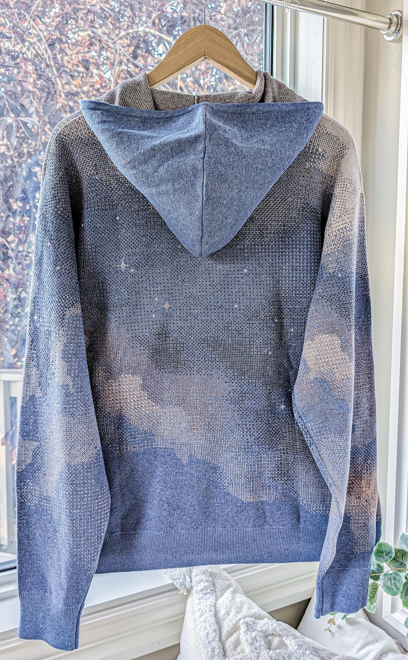 K&ouml;nnte beinhalten: Ein blauer Strick-Kapuzenpullover mit einem dezenten, gesprenkelten Muster aus hellen und dunklen T&ouml;nen, das an einen sternenklaren Nachthimmel oder Wolken erinnert. Das Kleidungsst&uuml;ck hat eine Kapuze, lange &Auml;rmel und einen gerippten Saum.