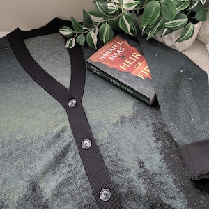 May include: A black and green cardigan with a black button closure. The cardigan has a subtle pattern of small dots. A book titled "Heir of Fire" by Sarah J. Maas is resting on top of the cardigan.