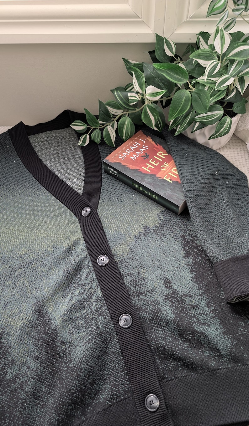May include: A black and green cardigan with a black button closure. The cardigan has a subtle pattern of small dots. A book titled "Heir of Fire" by Sarah J. Maas is resting on top of the cardigan.