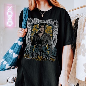 May include: Black t-shirt featuring a graphic print of a man with a sword and a dragon design. The text on the shirt reads "Haden Rierson, Rierson House, Sat Oct 10".