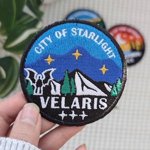 May include: Embroidered patch with the text "CITY OF STARLIGHT" in white on a blue background. The patch features a mountain scene with trees, stars, and the word "VELARIS". The patch is round with a black border.