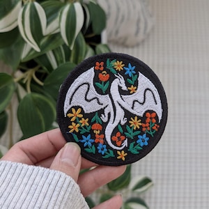 May include: Round embroidered patch featuring a white dragon with outstretched wings, surrounded by colorful flowers in orange, yellow, and blue. The patch has a black border and is likely for clothing or accessories.