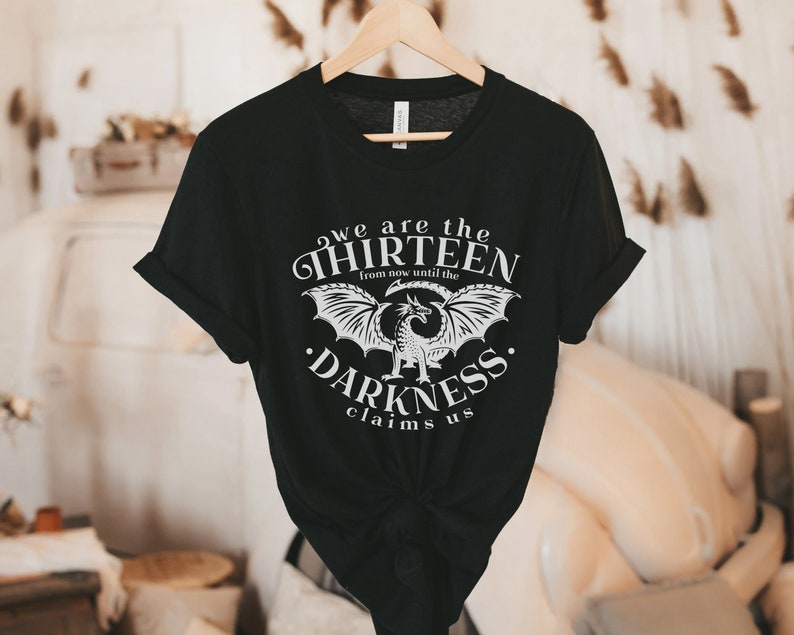 May include: Black t-shirt with a white graphic design featuring a dragon and the text "we are the THIRTEEN from now until the DARKNESS claims us." The shirt is hanging on a wooden hanger.