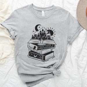 May include: A gray t-shirt with a black and white graphic of a stack of books with a castle and forest scene on the top book. The text on the shirt reads "I have lived a thousand lives."